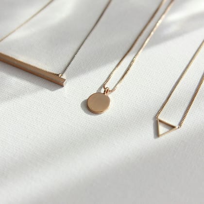Three fine rose gold necklaces with a bar, a disc and a triangle, on white